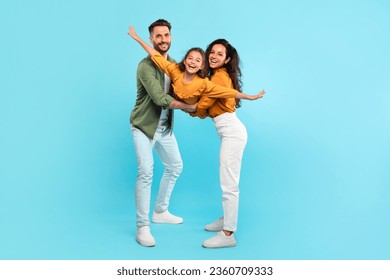 Excited parents playing with daughter holding her in arms posing on blue background, full length, family having fun together, girl spreading hands like plane