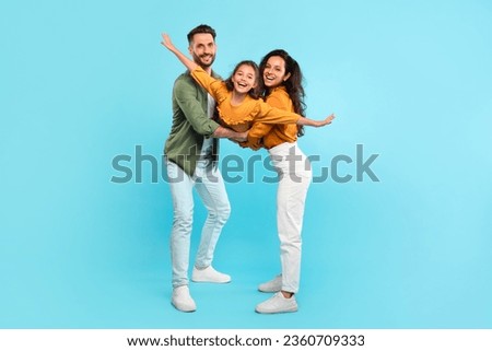 Excited parents playing with daughter holding her in arms posing on blue background, full length, family having fun together, girl spreading hands like plane