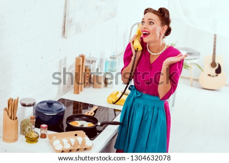 Excited pin up girl talkin on vintage yellow phone while frying eggs