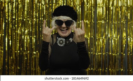 Excited Positive Senior Old Woman Showing Rock And Roll Gesture And Sincerely Smiling, Lady Enjoying On Concert, Activity. Elderly Stylish Grandma Showing Tongue On Golden Background