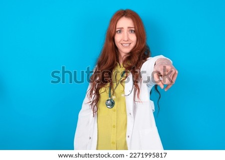 Excited positive young red-haired doctor woman wearing medical uniform over blue studio background points index finger directly at you, sees something very funny. Wow, amazing