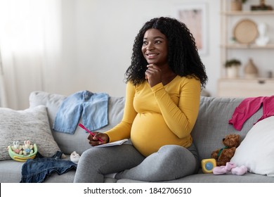 Excited Pregnant Black Woman Sitting On Sofa With Notebook And Pen, Making Shopping List Before Delivery, Free Space. Pensive African American Mother Getting Ready For Baby Coming, Home Interior