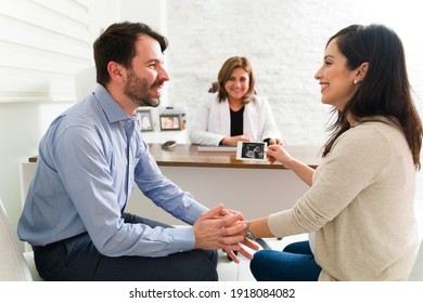 Excited pregnant couple holding the print ultrasound of their baby while sitting with their female gynecologist at her desk. Happy pregnant woman and man just found out the sex of their baby