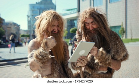 Excited prehistoric bushmen of hunter-gatherers covered in fur browsing internet on tablet discovering technology in modern city. Adaptation concept. - Shutterstock ID 1734279020