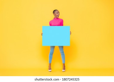 Excited pretty African American woman holding blue paper board with empty space for text on yellow isolated studio background