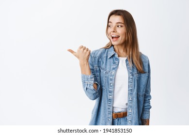 Excited Pretty Millennial Girl Turn Head And Pointing Thumb Left, Checking Out Good Promo Offer, Demonstrating Advertisement On Copy Space, White Background