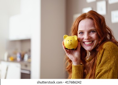 Aufgeregt mit einer hübschen, roten Kopfweibchen mit langen Haaren und gelbem Pullover, die ein süßes, gelbes Sparschwein in der Nähe ihrer Wange hält