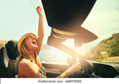 Excited pretty woman with hat and sunglasses reaching her fist in air as if to celebrate in convertible top automobile