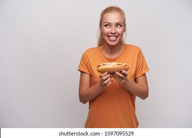 Excited pretty young long haired blonde lady with hot dog in raised hands looking aside with agitated smile and foretasting delicious dinner, standing over white background in orange t-shirt