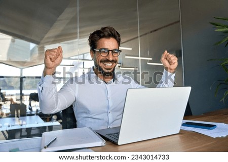 Excited professional young Latin business man celebrating success raising fist happy about financial bank market growth or reward work results, corporate goals achievement looking at laptop in office.