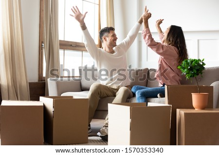 Excited proud young husband and wife renters tenants raise hold hands celebrate moving day buying new house, happy couple first time home buyers sit on sofa in own flat with boxes, relocation concept