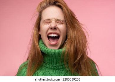 Excited redhead female in green sweater closing eyes and yelling against pink background