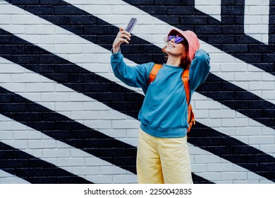 Excited redhead woman taking a selfie photo outdoors. Emotional hipster fashion woman in bright clothes, heart shaped glasses, bucket hat taking selfie photo on the phone camera. Street fashion. - Powered by Shutterstock