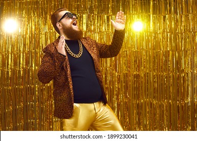 Excited Rich Red-haired Young Man In Neck Chain And Cool Glasses Having Fun At Glamour Night Club Disco Party. Happy Fashionable Bearded Showman Singing And Dancing On Stage With Golden Background