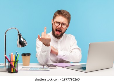 Excited Rude Man With Beard Office Worker Showing Middle Finger Looking At Camera Sitting At Workplace, Uncultured Gesture, Disrespect And Negativity. Indoor Studio Shot Isolated On Blue Background