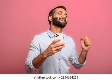Excited satisfied multiracial man holding smartphone and rejoicing isolated on pink. Surprised emotional lucky Indian man with received good message, news, won the lottery