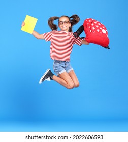 Excited schoolgirl in glasses captured in mid air, jumping up from happiness and joy with backpack and exercise book in hands, excited and ready for school, isolated over blue studio background