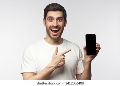 Excited Screaming WOW Young Man In White T-shirt Presenting Smartphone And Pointing With Finger At Blank Black Screen, Isolated On Gray Background