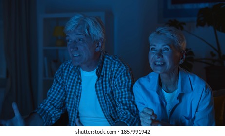 Excited Senior Couple Relax On Couch Watching Sports Game On Tv. Portrait Of Aged Husband And Wife Watching Football And Celebrating Victory Of Favorite Team At Home In Evening