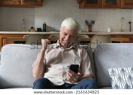 Excited senior grandpa sit on couch scream raise fist up in winner gesture read great pleasant news in email on cell screen. Joyful mature man celebrate success get victory in game on mobile phone