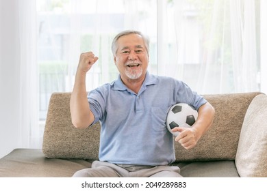 Excited Senior Man , Old Man Feel Happy Holding Football Soccer Ball Prepare For Cheer Team Favorite  On Couch Sofa At Home , Lifestyle Asian Senior Good Health Concept