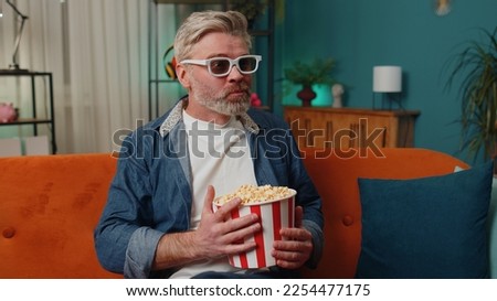 Excited senior mature man sits on couch eating popcorn snacks and watching interesting TV serial sport game, film, online social media movie content at home. Elderly guy enjoying evening entertainment