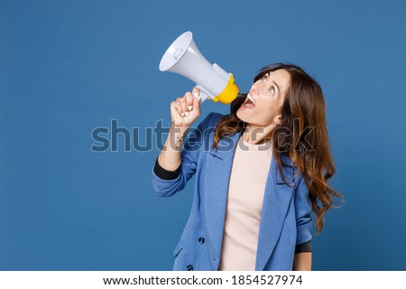 Excited shocked cheerful funny attractive young brunette woman 20s wearing basic jacket screaming in megaphone looking aside up isolated on bright blue colour wall background studio portrait