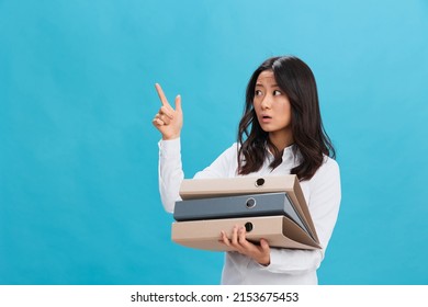 Excited shocked cute Asian businesswoman in classic office dress code holds a lot of folders point finger up posing isolated on blue studio background. Cool business offer. Too much work tasks concept