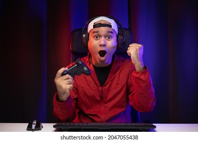 Excited and shocked face of Asian gamer with headphone holding joystick playing video game online sitting on chair at living room. Indian professional gamer streaming on social playing game very fun