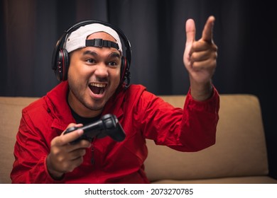 Excited And Shocked Face Of Asian Gamer With Headphone Holding Joystick Playing Video Game Online Sitting On Sofa At Living Room. Indian Professional Gamer Streaming On Social Playing Game Very Fun