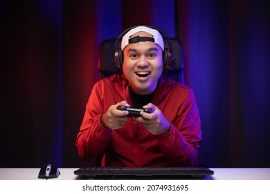 Excited And Shocked Face Of Asian Gamer With Headphone Holding Joystick Playing Video Game Online Sitting On Chair At Living Room. Indian Professional Gamer Streaming On Social Playing Game Very Fun