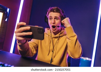 Excited And Shocked Gamer With Headset Playing Video Mobile Game Online On His Smartphone, Sitting On Gaming Chair In Neon Room. Professional Gamer Streaming The Game, Having Fun, Rejoices In Victory