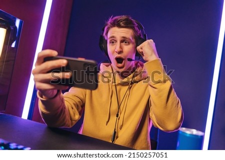 Excited and shocked gamer with headset playing video mobile game online on his smartphone, sitting on gaming chair in neon room. Professional gamer streaming the game, having fun, rejoices in victory