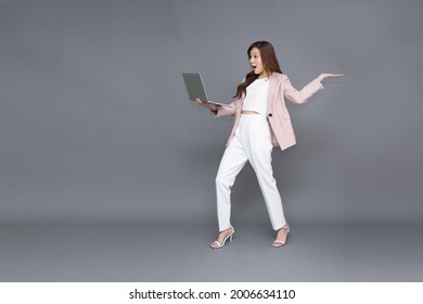 Excited Shocked Pretty Young Asian Business Woman In Pink Suit Holding Laptop Computer Or Notebook And Feeling Say Wow Isolated On Grey Background, Full Length People Composition