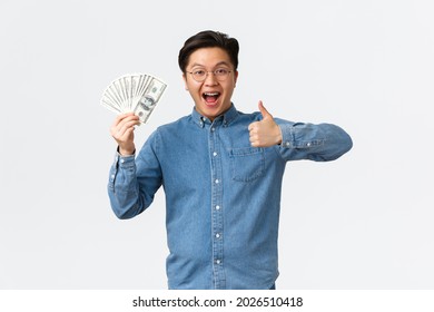 Excited Smiling Asian Man With Braces And Glasses, Showing Thumbs-up And Waving Money, Receive Paycheck, Freelancer Got Job With Good Payment, Holding Cash And Looking Satisfied