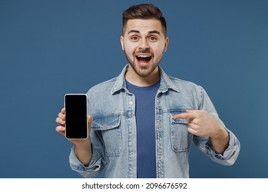 Excited smiling overjoyed young brunet man 20s wears denim jacket hold in hand use mobile cell phone with blank screen workspace area pointing on it isolated on dark blue background studio portrait. - Powered by Shutterstock