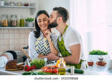 Excited smiling young couple in love making a super healthy vegan salad with many vegetables in the kitchen and husband kissing his wife