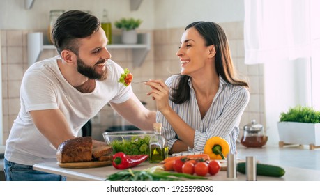 Excited smiling young couple in love making a super healthy vegan salad with many vegetables in the kitchen and man testing it from a girl's hands