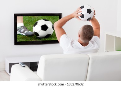 Excited Soccer Fan Watching A Game On Television Holding A Soccer Ball Above His Head As He Sits On A Comfortable Sofa In His Living Room