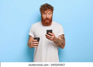 Excited speechless man stares at cellular, gasps from wonder, receives message, finds out about raising salary, holds takeaway coffee, has tattoo on arm, wears white t shirt, ginger thick beard - Shutterstock ID 1493293457