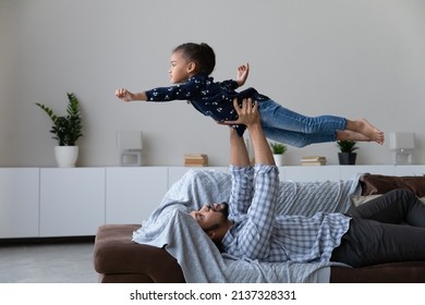 Excited Strong Young Black Dad Lying On Couch, Lifting Kid Up In Air, Holding Little Daughter Girl Playing Flying Airplane With Open Hands, Doing Power Support Exercise, Acroyoga