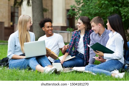 Excited students resting in campus outdoors, sitting on grass, chatting and using laptop