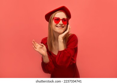 Excited stylish happy laughing girl holding hand on face or keeping hand on cheek, wearing beret headwear, red dress with heart glasses and looking up isolated on colored pink background.