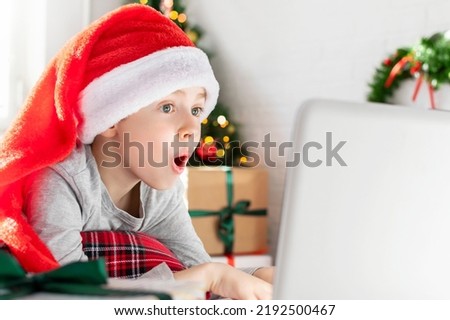 Excited surprised boy in a red Santa hat looks at the laptop screen. A shocked child lies at home in front of the Christmas tree. Merry Christmas online shopping, sale season.