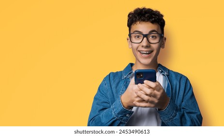 Excited surprised shocked astonished happy curly haired funny young man wear braces glasses spectacles open mouth hold typing cell phone cellular smartphone cellphone isolated yellow background - Powered by Shutterstock