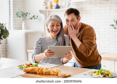 Excited Surprised Shocked Mature Middle-aged Couple Family Wife And Husband Reading News, Watching Movie Searching Fro Sales On Digital Tablet During Breakfast In The Kitchen Together At Home