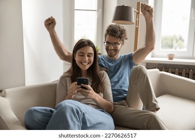 Excited surprised successful young couple using mobile phone together, resting on sofa at home, making winner hand gesture, throwing fists up. celebrating success, achieve, luck, laughing, shouting