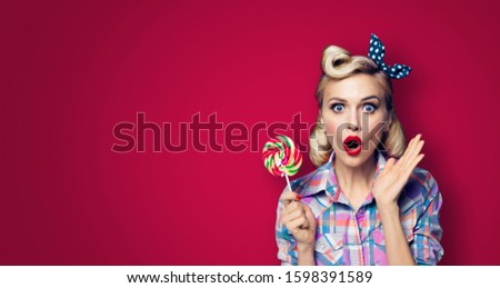 Excited surprised woman with lollipop. Girl pin up with open mouth. Blond model at retro fashion and vintage concept. Dark red color background. Copy space for some advertise slogan, sign or text. 
