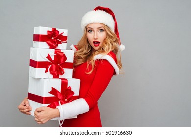 Excited surprised woman in red santa claus outfit holding stack presents isolated on the gray background
