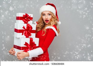 Excited surprised woman in red santa claus outfit holding stack presents isolated on the gray background - Powered by Shutterstock